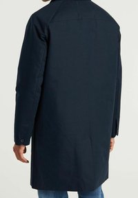 Man som bär en lång, mörk marinblå kappa sedd bakifrån, med knäppta manschetter och blå jeans synliga under kappans nederkant.