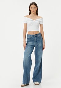 Hvid off-the-shoulder crop top med twist detalje, parret med lyseblå vide ben denim jeans og beige sneakers. Glatte teksturer og afslappet pasform.