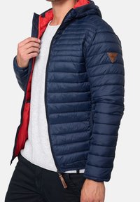 Navy blue puffy jacket with red interior, zip closure, quilted texture, attached hood, and a brown leather logo patch on the sleeve.