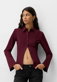 Burgundy skjorta med krage, långa utsvängda ärmar och knäppning fram, utformad för att framhäva midjan med en figurnära silhuett.