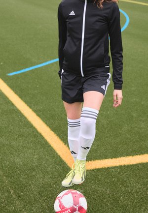 Person iført sort Adidas sportstøj kontrollerer en rød og hvid fodbold på en grøn bane markeret med gule og blå linjer.