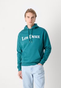 Teal hooded sweatshirt with white "Les Deux" text, kangaroo pocket, and ribbed cuffs. Paired with light blue jeans.