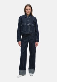 Czarny dżinsowy komplet składający się z dwóch części z prążkami. Krótka kurtka z przednimi kieszeniami i srebrnymi guzikami. Spodnie z szerokimi nogawkami zakończone mankietami. Białe obuwie.