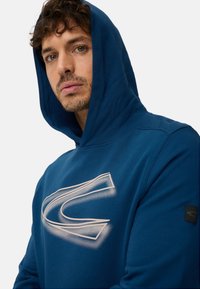 Blå hættetrøje med et stort hvidt grafisk logo trykt på brystet. Har snøre i hætten og ribkant ved ærmerne for en tæt pasform.