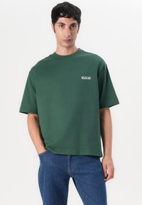 Tricou verde oversized cu guler rotund, mâneci scurte și un detaliu logo alb pe față; materialul pare neted și texturat.