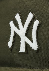 Dark green fabric with a raised white felt NY logo. The design features textured stitching, creating a pronounced visual contrast.