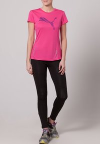 Camiseta deportiva de color rosa brillante con logotipo azul de Puma, combinada con leggins negros con paneles laterales, usados con zapatillas de correr grises y amarillas.