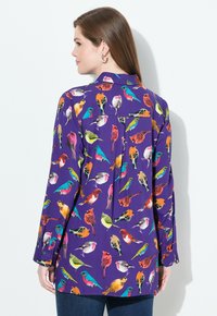 Blusa viola con motivi di uccelli colorati, maniche lunghe, polsini con bottoni e dettagli con yoke sul retro. Tessuto leggero, vestibilità comoda.