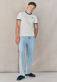 Biała koszulka z krótkim rękawem, niebieskim prążkowanym kołnierzem i logo na lewej piersi, zestawiona z jasnoniebieskimi luźnymi jeansami i czarnymi skórzanymi butami.
