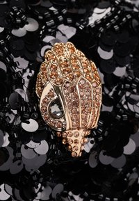 Spilla in tonalità oro a forma di mano, adornata con gemme marroni, su uno sfondo di paillettes nere. Texture e lucentezza ben definite.