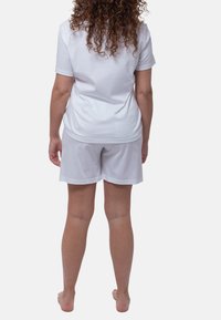 Nainen, jolla on kihara hius, seisoo paljain jaloin, selkä kameraan, päällä pelkkä valkoinen t-paita ja valkoiset shortsit yksinkertaista taustaa vasten.