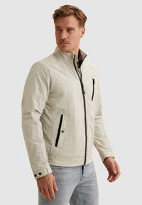 Homme portant une veste beige clair avec une fermeture éclair noire et des détails de poche, debout devant un fond uni, regardant vers la gauche.