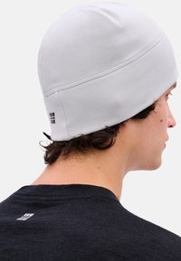 Witte gebreide muts met een gestructureerd patroon, voorzien van een logovlaggetje aan de achterkant. Draagt door een persoon met donker haar, gezien van achteren.