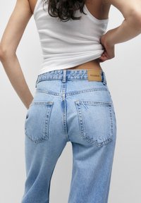 Mujer con jeans de mezclilla azul claro y top blanco, levantando la parte trasera de su top con una mano, de espaldas a la cámara.