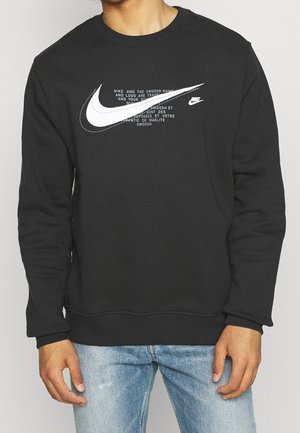 Sudadera negra con mangas largas, que presenta un gran logo blanco de Nike Swoosh y un diseño de texto en varios idiomas en la parte frontal.
