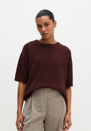Femme aux cheveux foncés portant un pull texturé marron foncé à manches courtes et un pantalon plissé taille haute gris clair sur un fond uni.