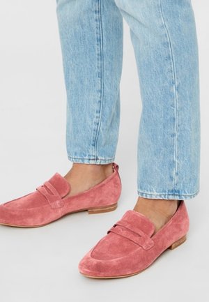 Voeten met roze suède loafers gecombineerd met lichtblauwe cropped jeans tegen een witte achtergrond.