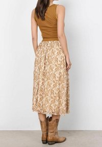 Ruskea istuva hihaton paita yhdistetty pitkään beigeen kukkaskettiin, jossa on abstrakti kuvio, ja se on täydennetty ruskeilla cowboy-kengillä.