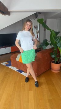 Νεαρή γυναίκα στέκεται σε σαλόνι φορώντας λευκό μπλουζάκι, πράσινη φούστα με σχέδια και μαύρα παπούτσια, με ένα μεγάλο φυτεμένο φυτό κοντά της.