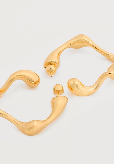 Ασύμμετρα σκουλαρίκια κρίκοι από χρυσό μέταλλο με ακανόνιστο σχήμα και κούμπωμα με καρφί, σε λευκό φόντο.