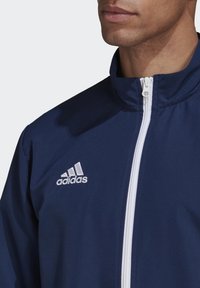 Navy lichtgewicht jack met hoge kraag, witte rits en wit Adidas-logo op de linkerkant van de borst. Gladde textuur en sportief ontwerp.