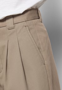 Close-up van beige geplooide broeken met zijzak en riemlussen, gedragen met een witte, ingestopte blouse.