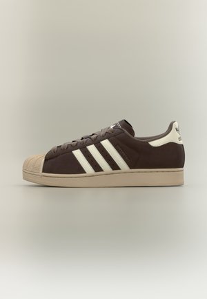 Brązowy sneaker Adidas Superstar z beżowym gumowym noskiem, kremową podeszwą, białymi trzema paskami i pętelką na pięcie, widok z boku na neutralnym tle.