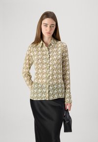 Camicia con bottoni in tessuto leggero, caratterizzata da un motivo geometrico giallo, marrone e bianco; abbinata a una gonna nera in satin e a una piccola borsa nera.