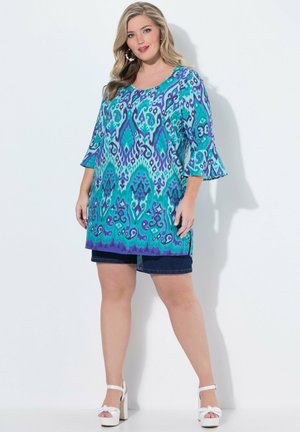 Kobieta nosząca niebieską i fioletową tunikę we wzory z rozkloszowanymi rękawami, ciemne krótkie spodenki, białe sandały na wysokim obcasie i duże kolczyki koła.