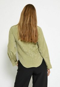 Camicia a maniche lunghe verde oliva chiaro con tessuto arricciato, polsini con bottoni, orlo arrotondato, indossata con pantaloni neri.