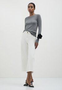 Grauer Langarmpullover mit gerippten Bündchen, kombiniert mit weißen weiten Hosen und schwarzen Slingback-Highheels mit dekorativen Akzenten.