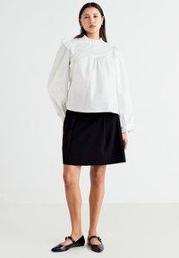Blouse blanche à manches longues avec un col et des poignets froncés, associée à une jupe noire en forme de A, portée avec des chaussures plates noires.