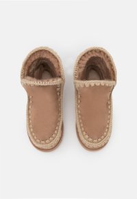 Pantofole in pelle marrone chiaro con rivestimento morbido marrone scuro, rifinitura intrecciata attorno all'apertura e punta arrotondata. Suola piatta con una leggera texture.