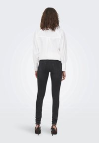 Camisa blanca de botones con mangas abullonadas y yugo en la parte posterior, combinada con unos jeans ajustados negros y zapatos de tacón alto. Diseño simple y limpio.