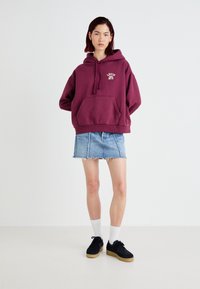 Burgundy hoodie med framficka och dragsko på huvan, tillsammans med en ljus denimkjol. Svarta skor och vita strumpor fulländar outfiten.