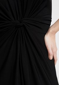 Musta mekko, jossa solmittu selkäosa, ryppyistä kangasta luomassa tekstuuria ja sileä, joustava materiaali. Korostaa vartalon muotoja ja istuvuutta.
