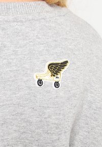 Sweatshirt gris avec un patch brodé proéminent représentant un roller avec des ailes noires, détaillé en fil doré et noir.