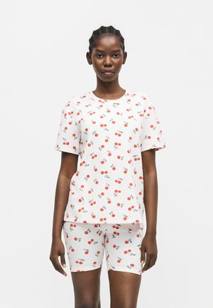 Mladá žena nosí biele tričko s krátkym rukávom a zodpovedajúce krátke nohavice potlačené červenými vzormi čerešní, stojí pred jednoduchým pozadím.