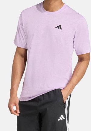 Muž nosící světle fialové tričko s krátkým rukávem a černé sportovní šortky s bílým logem a bílými pruhy na stranách, ruka v kapse.