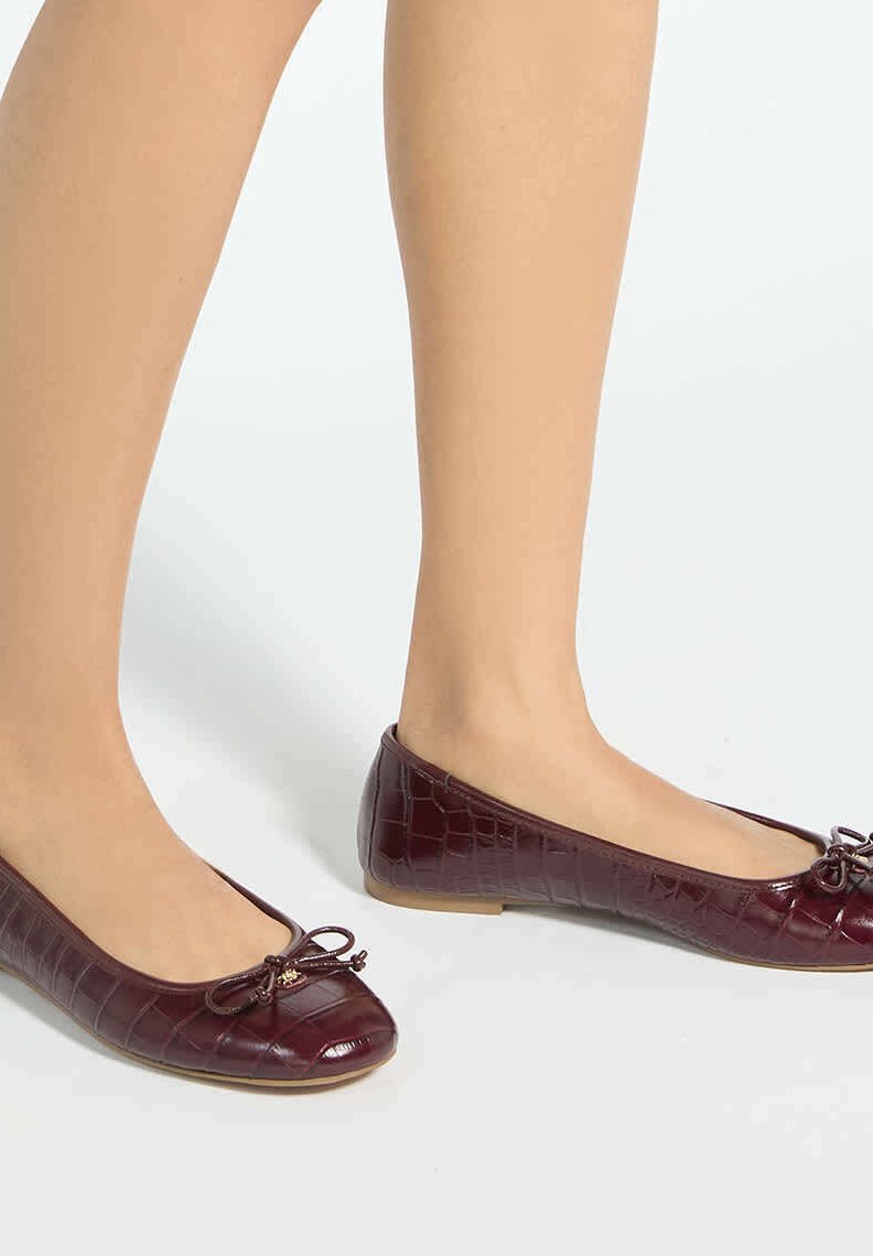 Jambes nues portant des ballerines en cuir texturé crocodile bordeaux foncé avec petits nœuds sur le dessus, debout sur une surface claire.