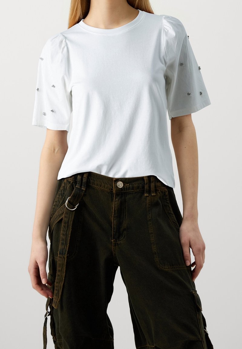 Femme portant une chemise blanche à manches courtes avec de petites décorations sur les manches et un pantalon cargo sombre et ample avec des sangles.