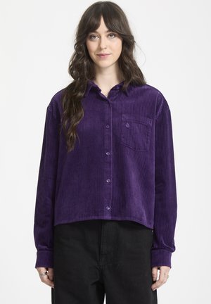 Lilla kordfløjl skjorte med knaplukning foran, lange ærmer og en enkelt brystlomme. Stoffet har en vertikal ribbet tekstur.