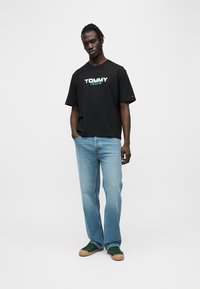 Μαύρο βαμβακερό μπλουζάκι με το λογότυπο "TOMMY JEANS" σε λευκό και πράσινο, συνδυασμένο με τζιν παντελόνι ανοιχτού μπλε και σκούρα πράσινα αθλητικά παπούτσια.