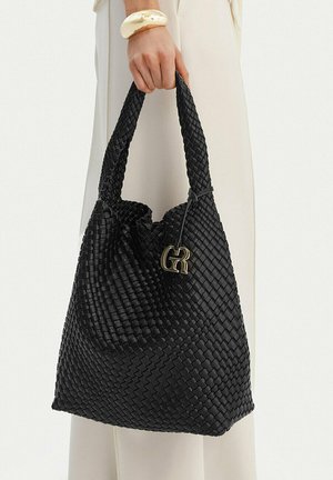 Ruka držící velkou černou tkanou koženou kabelku s očkovou rukojetí a zlatavým ozdobným přívěskem.