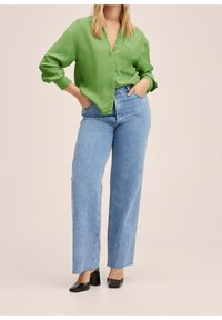 Femme portant une blouse verte à manches longues, un jean taille haute bleu clair et des chaussures noires à talons carrés, debout avec les mains dans les poches.