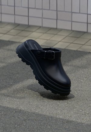 Czarny klapek wsuwany na grubej, protektorowej podeszwie z regulowanym paskiem, ustawiony nad asfaltem na tle ściany z kafelków.