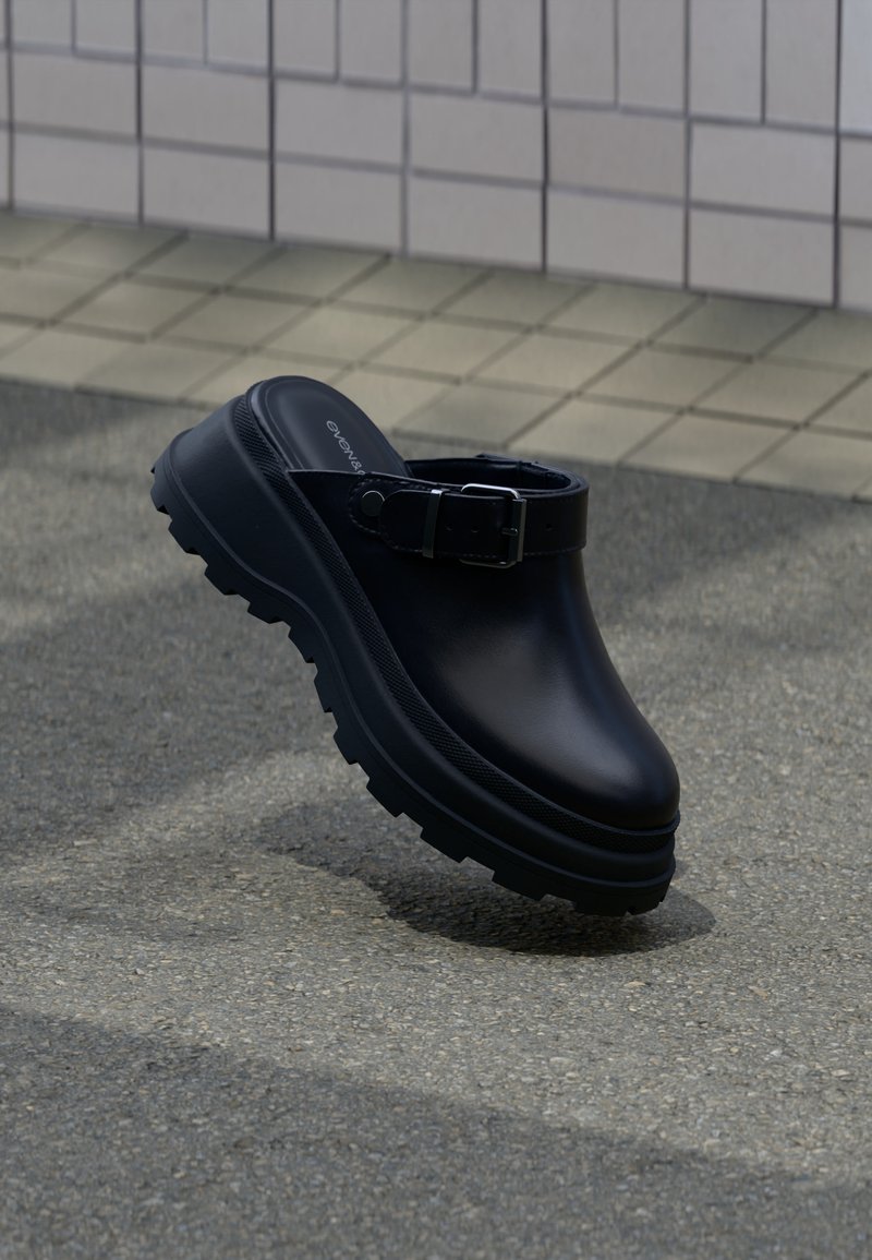 Sabot noir à enfiler avec semelle épaisse crantée et bride réglable, posé sur un sol en asphalte avec un mur carrelé en arrière-plan.