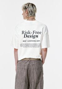 Hvid oversized T-shirt med sort tekst på ryggen. Parret med strukturerede, taupe-farvede bukser. Enkel og minimalistisk design.