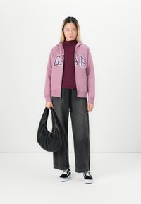 Lichtpaarse zip-up hoodie met "GAP" logo, bordeaux turtleneck, zwarte wijdlopende jeans en zwarte hobo tas. Witte sneakers met zwarte accenten.