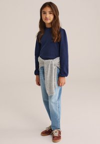 Jūras spēku garās piedurknes topiņš, pelēks džemperis sasiets ap vidukli, gaiši zili džinsi un brūni zamšāda sporta apavi ar sarkaniem akcentiem.